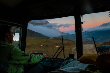 Young Caucasian man with glasses drives a vehicle through the mountainous landscape at sunset. Clouds hover over the hills.