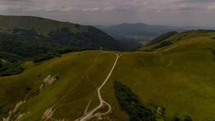 Aerial view of the Caucasus mountains with rolling green hills and a winding dirt road. The landscape is expansive and serene, showcasing natural beauty.