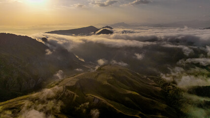 Aerial view of the Caucasus mountains with rolling hills and clouds. The sun sets in the background, casting a warm glow over the landscape.