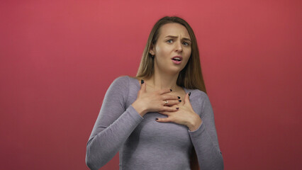 Obraz premium Woman expressing fear with hands pressed to chest against an isolated red background, showcasing a young caucasian woman's emotion and reaction vividly.