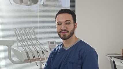 Chest up portrait of professional young Middle Eastern male dentist in scrubs smiling at camera while sitting on chair in contemporary dental office supplied with modern equipment
