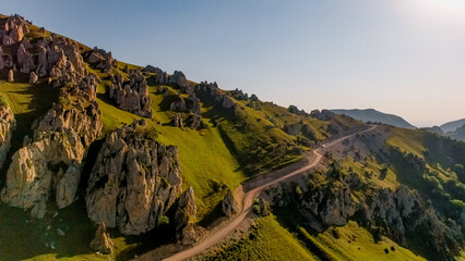 Aerial view of lush green hills and rocky formations in the Caucasus region. A winding road cuts through the landscape under a clear blue sky.