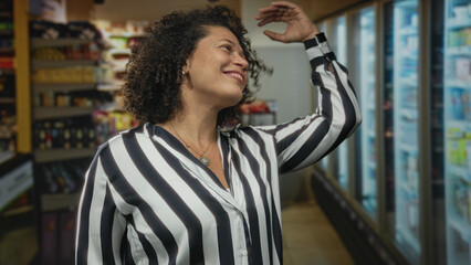 Woman touches hair and smiles in a supermarket aisle with glass refrigerator doors and stocked shelves; joy.