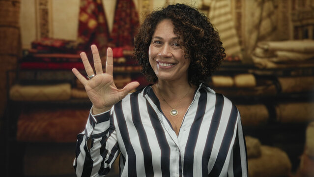 Woman counts on her fingers and holds up an open hand showing five among stacked carpet rolls in a shop interior; friendly greeting.