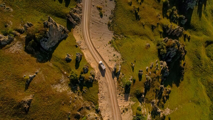 Aerial view of a winding dirt road through green hills and rocky formations in the Caucasus region. A vehicle is seen traveling along the road.