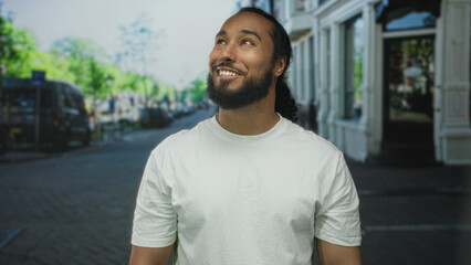 Fototapeta premium Man in white t-shirt smiling and looking up on a city street with storefronts and parked cars visible; optimism urban calm.