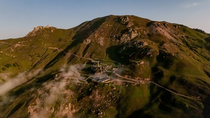 Aerial view of a mountainous landscape in the Caucasus region. Green hills and rocky terrain are visible, with a small settlement nestled among the peaks.