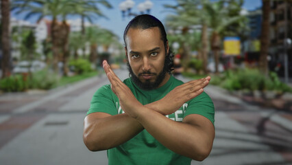 Fototapeta premium Man with crossed arms forming an x gesture, hands and forearms visible, wearing a green shirt while standing on a sunny street lined with palm trees; rejection protest.