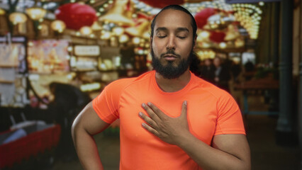 Obraz premium Man touching his hand to chest in street market with orange tshirt, lanterns and festive lights; serenity gratitude.