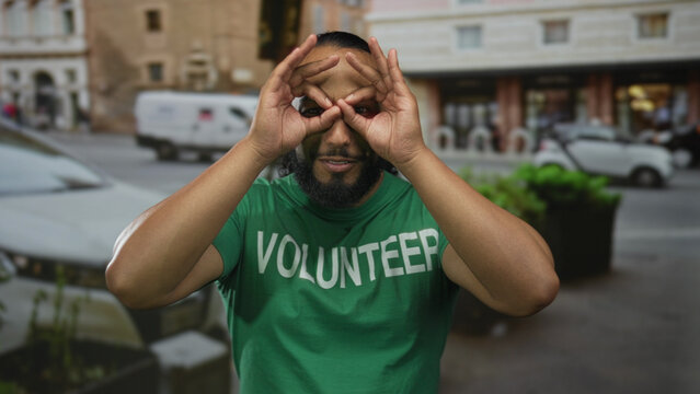 Man volunteer in a green shirt making binocular hand gesture over his eyes and smiling on a street; community service joy. - Powered by Adobe