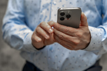 A man in a blue shirt and elegant trousers stands against a concrete wall with a cell phone in his hand