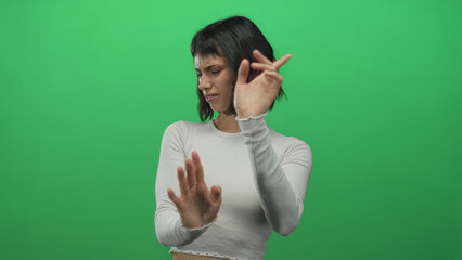 Young hispanic woman grimaces and raises open palms for stop gesture in front of green screen in studio; disgust.