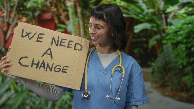 Woman nurse in blue scrubs holds cardboard sign we need a change among lush greenery in park; change activism determination.