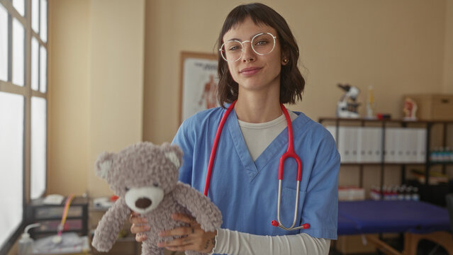 Woman doctor in blue scrubs holds teddy bear inside building with stethoscope around neck; compassion.