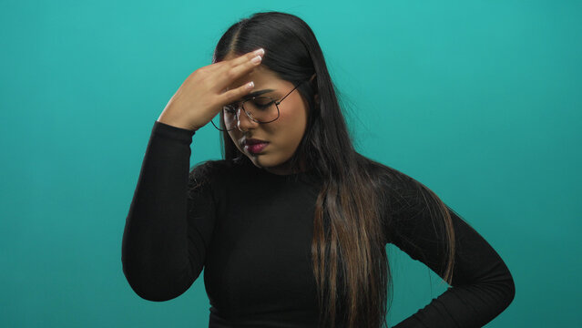 Young hispanic woman in black top looking stressed with hand on forehead against an isolated green background, displaying frustration or contemplation.