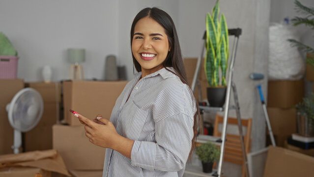 Woman texting on smartphone in new living room setting with moving boxes baring a thoughtful yet confident expression amidst an array of unpacked household items.