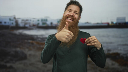 Man gives thumbs up while smiling broadly and holding a red heart cutout at a rocky shoreline beach scene behind him; love.