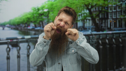 Man with beard making mock crying gesture with clenched fists near eyes on canal street bridge; playful mockery.