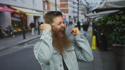 Young caucasian man with long beard wearing denim jacket raising clenched fists on city street;...