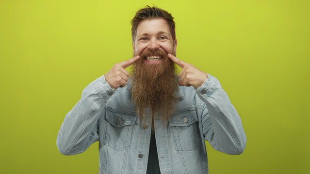 Man with long beard wearing denim jacket and black shirt points finger to teeth in a vibrant green studio; happiness.