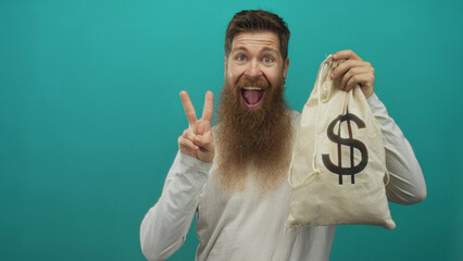 Bearded man holding large money bag and making peace sign in teal studio; wealth triumph...