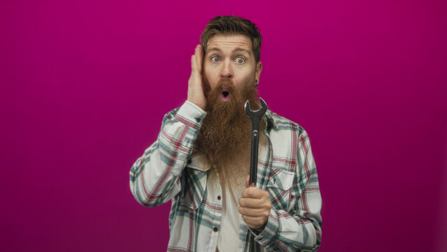 Man holds wrench with open mouth and raises hand to cheek in studio against magenta backdrop; surprise. - Powered by Adobe