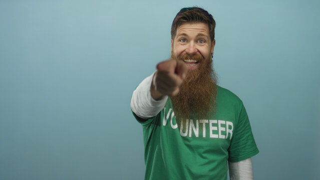 Man wearing volunteer shirt smiling and pointing finger to camera in blue studio; invitation motivation. - Powered by Adobe