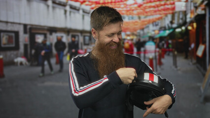 Bearded man pointing finger to camera while holding helmet under hanging lanterns on a busy street;...