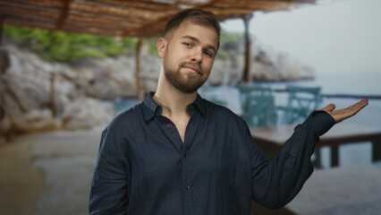 Man with open palm gesture at seaside building terrace, hand raised palm up toward ocean and rocky shore; serenity.