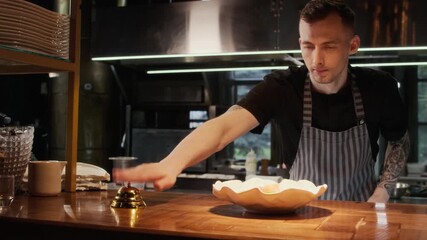 Medium shot of professional chef carefully placing finished dish on pass and ringing bell to be served to customers in restaurant kitchen, copy space