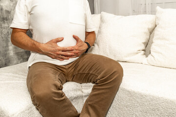 A young man in brown long pants sits on a white sofa in the living room, holding his stomach,