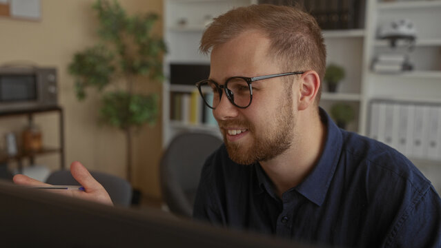Man wearing glasses and beard holding smartphone and smiling while gesturing with hand in an office building; concentration.