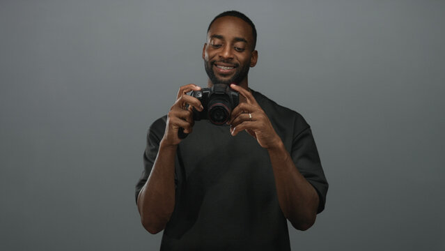 Man holding dslr camera, smiling and adjusting lens with gold chain visible in gray studio; confidence creativity inspiration.