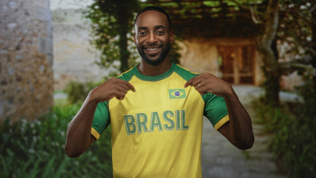 Man points to brasil jersey and flag patch with both hands at building entrance while smiling and showing braces; team pride.