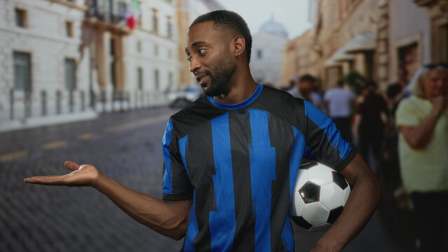 Man gives thumbs up while holding a soccer ball on a cobbled street with blurred supporters and buildings behind him; team support pride.