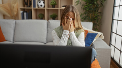 Blonde woman covers her face with both hands while seated on a sofa watching a screen in a living room; sadness.