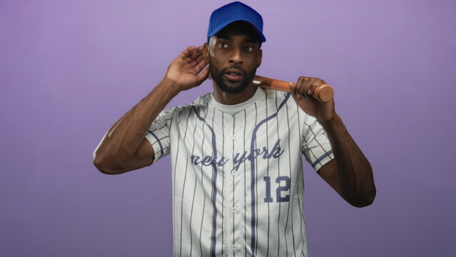 Man baseball player wearing blue cap and pinstripe jersey numbered 12 holds wooden bat across his shoulders with both hands in studio with purple backdrop; determination practice.