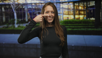 Woman pointing finger to nose while smiling and standing on a city sidewalk in front of a glass...