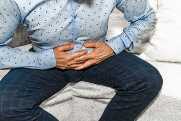 A man in elegant trousers and a long blue shirt sits on a white sofa, holding his aching stomach, hands on his stomach.