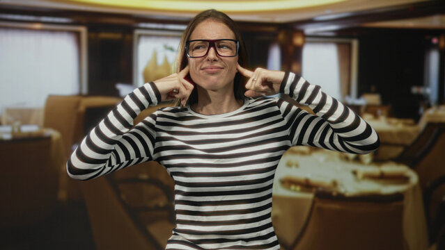 Woman covers ears with hands in building restaurant interior wearing striped shirt and glasses; stress frustration.