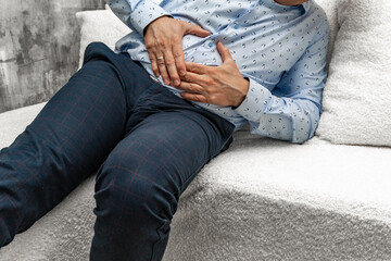 A man in elegant trousers and a long blue shirt sits on a white sofa, holding his aching stomach, hands on his stomach.
