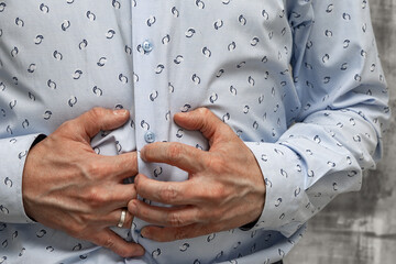 Close-up of a man's hands holding his aching stomach. A man wearing a blue long-sleeved shirt stands in a medical clinic