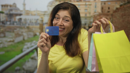 Woman smiling holds creditcard and colorful shopping bag by handles in hand at ancient roman building; happiness.