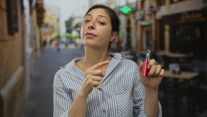Hispanic middle age woman holding red vape device and pointing finger on a busy city street; public...