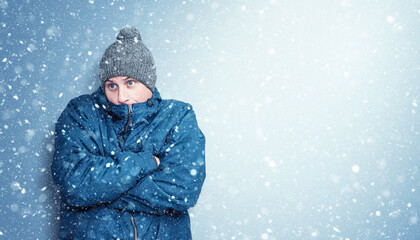 Cold man in a blue winter jacket and knit hat stands freezing in falling snow, arms crossed for warmth, with wide space on the right side for text or copy in a winter design.