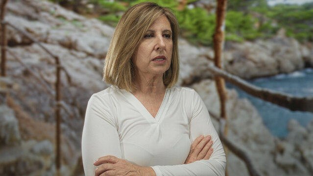 Woman standing with folded arms by seaside wooden railing on rocky coastline path under cloudy sky; concern.