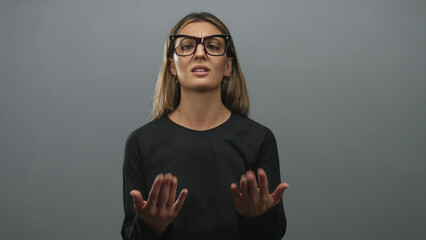 Woman wearing glasses rubbing fingers and pointing to open palm in studio with grey backdrop;...