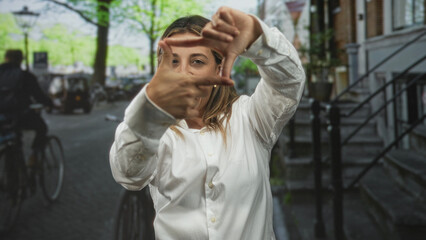 Obraz premium Young woman in white shirt framing face with hands on city street near stairs and lamppost; creative playful confidence.
