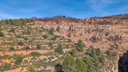 Aerial shot of dry Mediterranean mountain slopes with terraced terrain, rocky cliffs, and green trees in Spain.