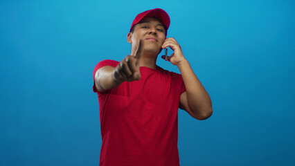 Man in red cap points finger at camera while holding smartphone in a bright blue studio; confidence friendly service.